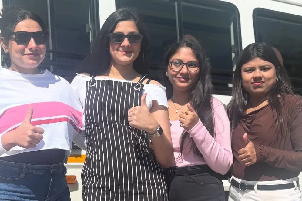 Four women standing shoulder to shoulder in front of a white minibus in bright sunshine, all four giving a thumbs-up to the camera.