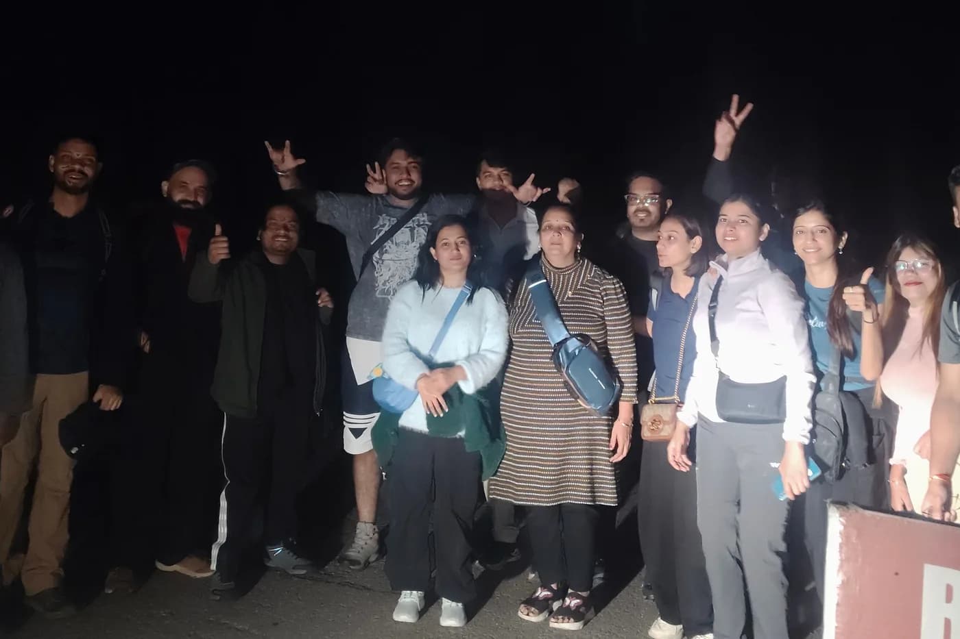 A group of about fourteen travellers in warm layers at a roadside stop after dark, several with hands raised to the camera.