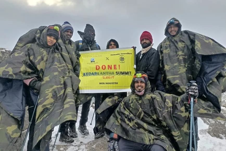 Eight trekkers in camouflage rain ponchos on the snow at the Kedarkantha summit, holding a banner reading 'Mistiii Tales x Nomadic Travellers — DONE! Kedarkantha Summit 12,500 ft'.